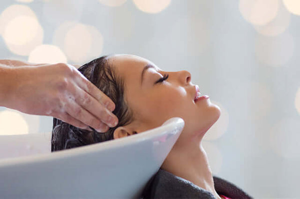 Woman lying back at a salon basin with her eyes closed as a stylist's hands work shampoo lather into her dark hair