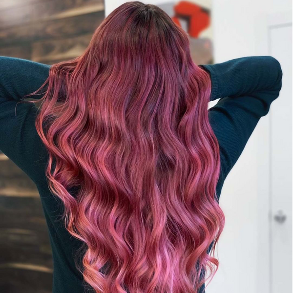 Back view of long magenta and pink balayage waves, arms raised, against a dark wood wall