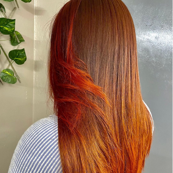 Back view of long copper hair with bright orange-red streaks, over a blue striped shirt by a gray wall