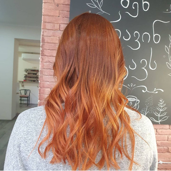 Long copper balayage waves from behind, deep auburn roots melting into bright ginger ends, in a salon with a chalkboard wall