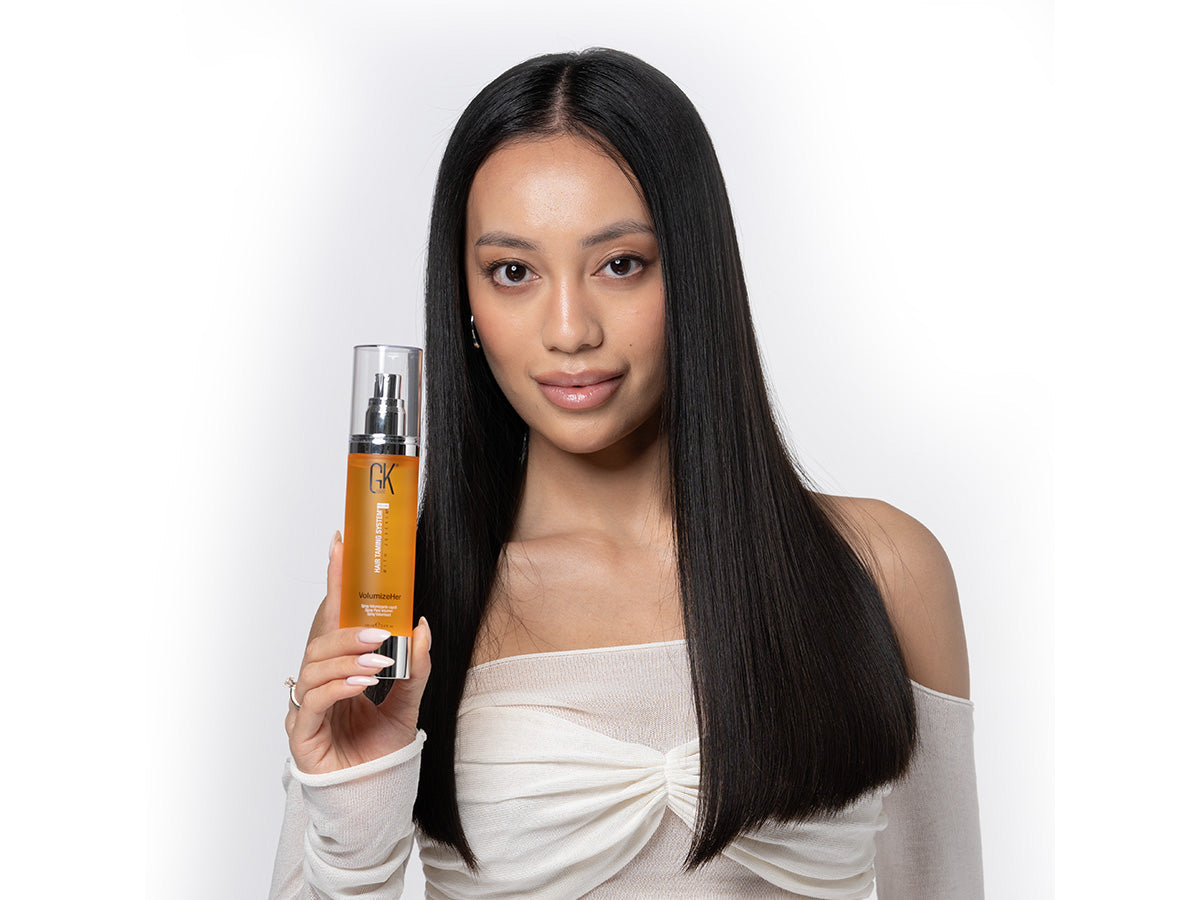 A person with straight, long hair holds a bottle of GK Hair Volumizing Spray, wearing a stylish off-shoulder top against a white background.