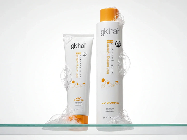GK Hair pH+ Shampoo 100 ml tube and 300 ml orange-capped bottle standing in soap foam on a glass shelf
