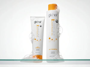 GK Hair pH+ Shampoo 100 ml tube and 300 ml orange-capped bottle standing in soap foam on a glass shelf