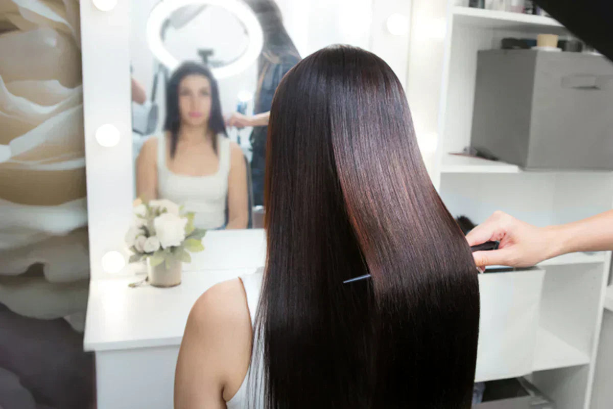 A woman with long, shiny hair is having her hair styled in a beauty salon, with a mirror and soft lighting in the background.