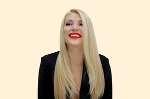 A woman with sleek, light blonde hair and red lipstick smiling confidently in a black outfit against a beige background.