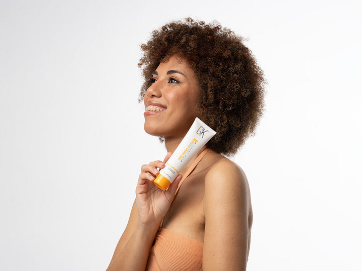 A lady with healthy short curly hair holding GK Hair ThermalStyleHer Cream
