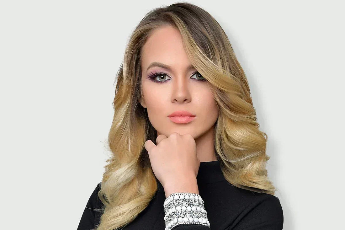 Woman with long, wavy blonde hair and pink lipstick, wearing a black top with jeweled cuffs, poses confidently against a plain background.
