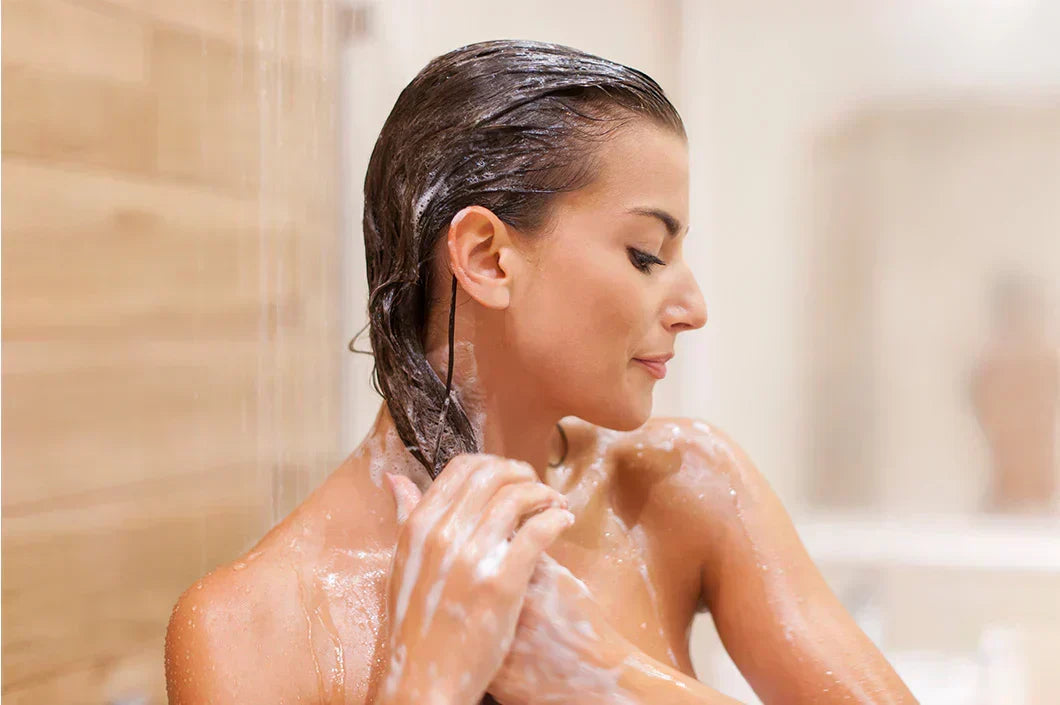 A person with closed eyes enjoys a shower, massaging shampoo into their hair. The warm, wooden background conveys a serene and relaxing atmosphere.