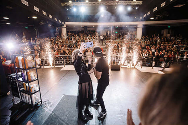 Stage finale at a GK Hair show: two presenters take a selfie between spark fountains as the audience applauds
