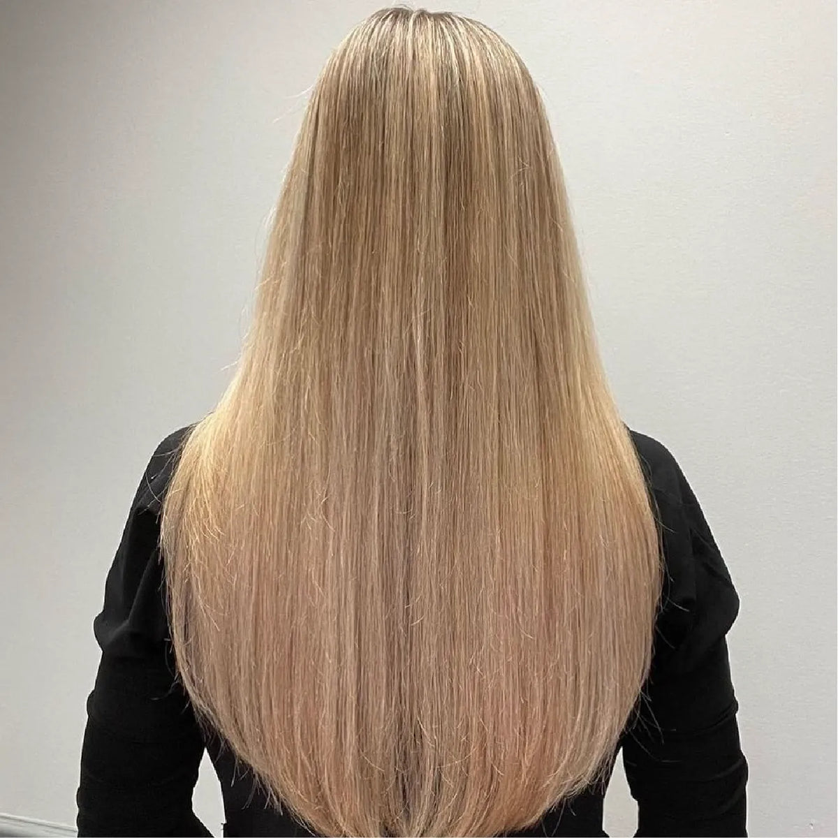 Back view of long, poker-straight golden blonde hair past the shoulders, black top, plain gray wall behind