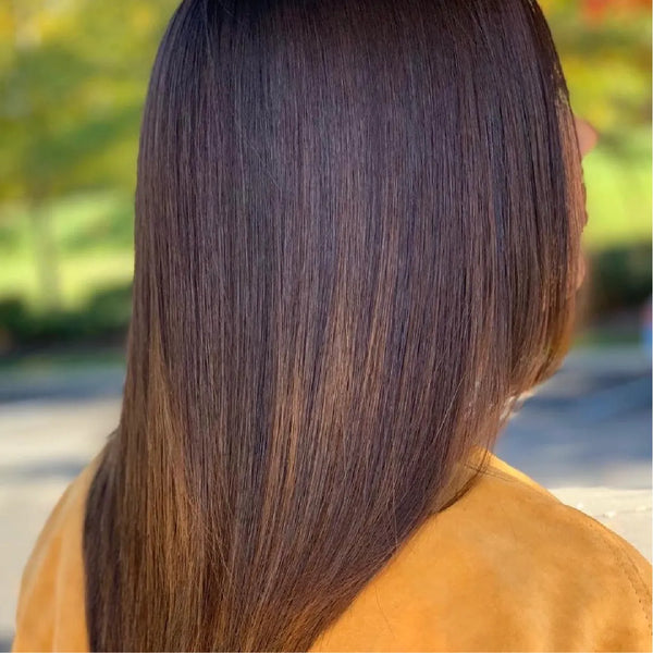 Back view of glossy dark brown hair with warm caramel ends over a mustard suede jacket, blurred trees behind