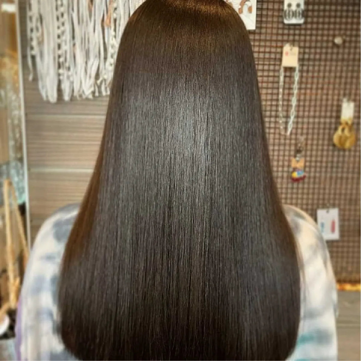 Back view of long, mirror-smooth dark brown hair on a woman in a tie-dye top, salon accessory display behind