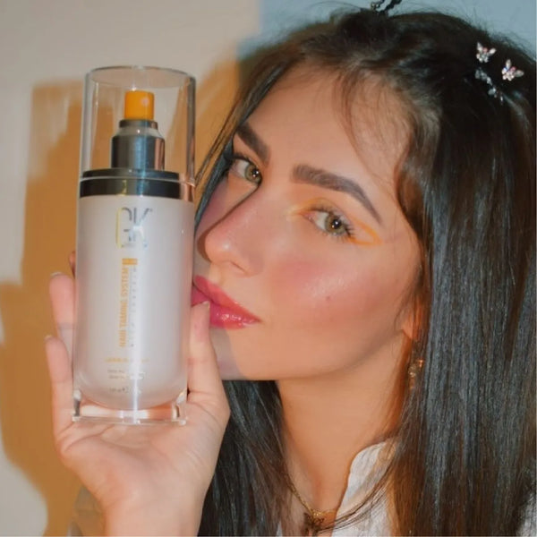 Dark-haired model in butterfly hair clips raises a frosted GK Hair pump bottle beside her cheek, orange and blue painted wall
