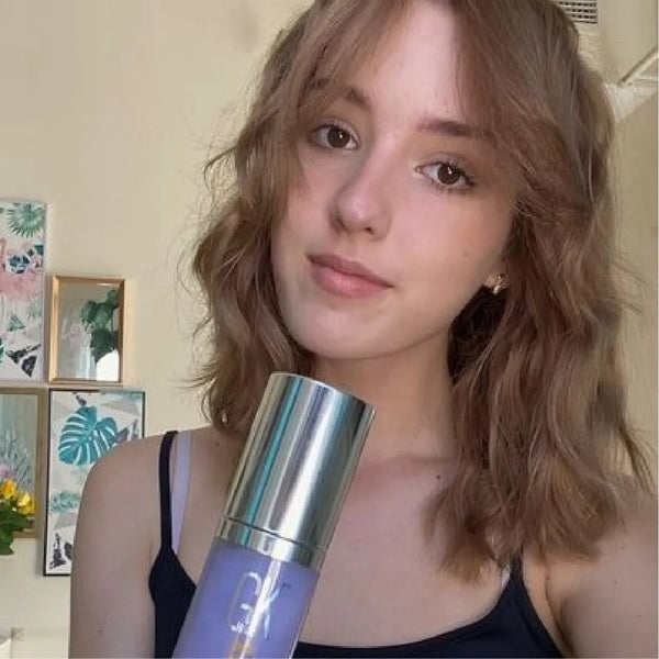 Wavy-haired model in a black tank top raises a lavender GK Hair bottle with a mirrored silver cap in front of a cream wall