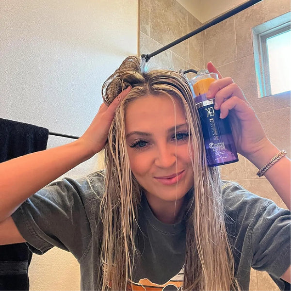Mirror selfie of a blonde with wet sectioned hair raising a GK Hair Leave-In Bombshell Cream bottle, tiled shower wall behind