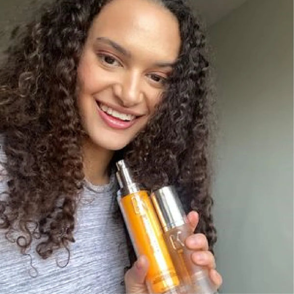 Curly-haired model in a heather gray top smiles, holding two slim bottles, one amber and one clear, up beside her shoulder