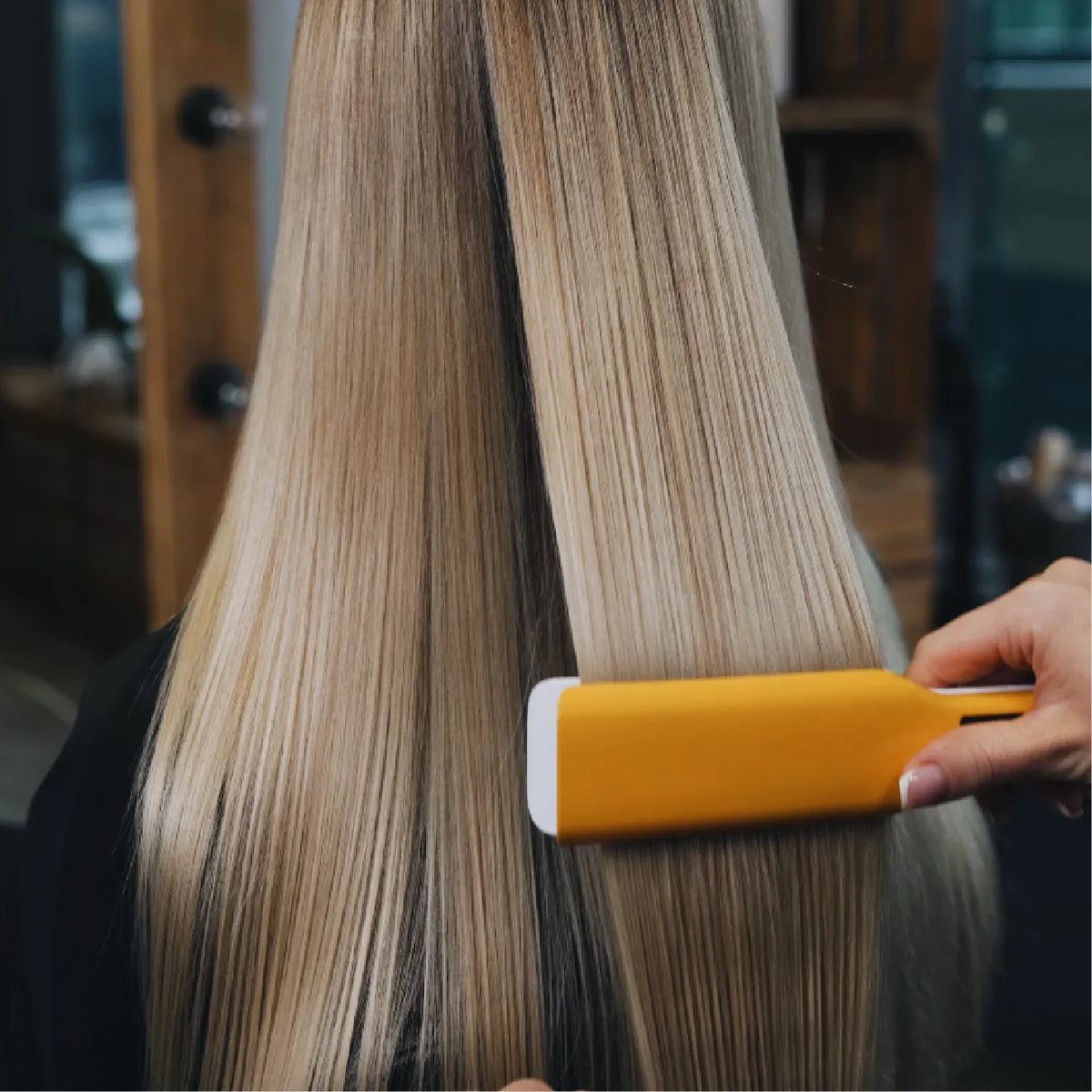 Long blonde hair seen from behind, sleek and straight, with a yellow and white flat iron pulled down a section in a salon