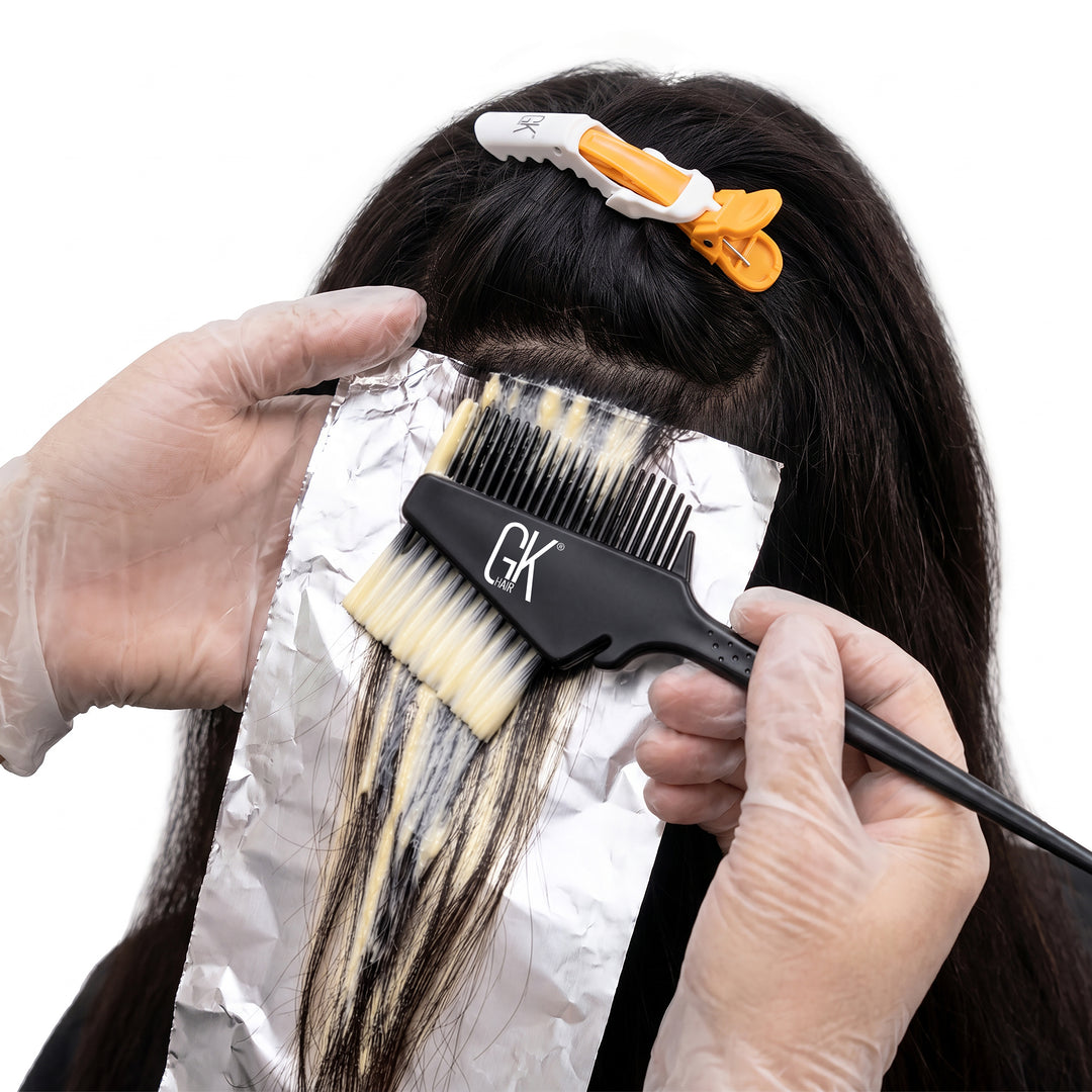 Gloved hands paint lightener onto a foiled section of dark hair with a black GK Hair brush-and-comb, orange clip above