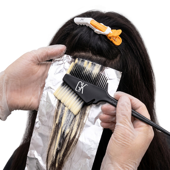 Gloved hands paint lightener onto a foiled section of dark hair with a black GK Hair brush-and-comb, orange clip above