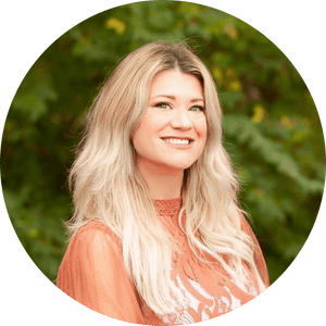 Circular headshot of a smiling woman with long wavy blonde balayage hair in a rust lace top, green foliage behind