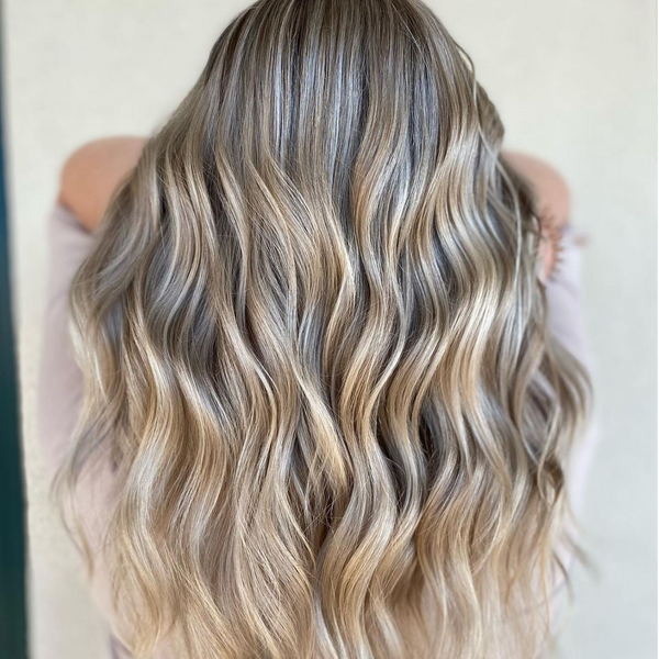 Back view of long beach waves in cool ash-blonde balayage over darker roots, against a cream wall
