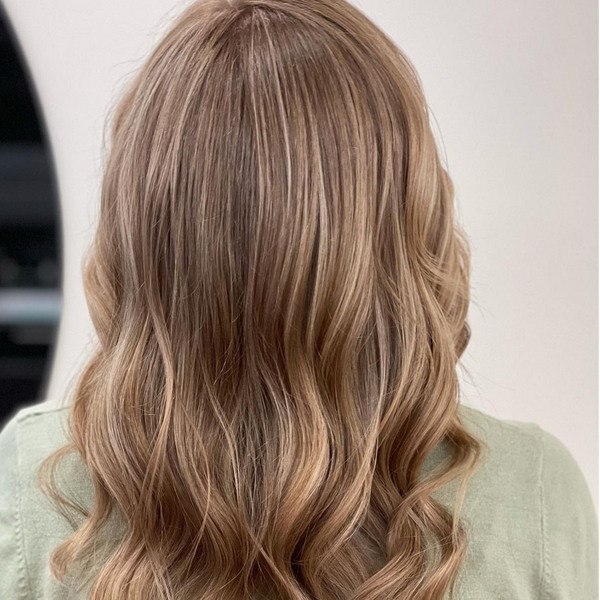 Back view of soft caramel-blonde waves over a sage green top, against a plain white salon wall