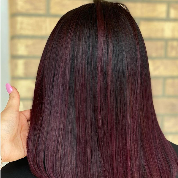 Back view of sleek black hair with deep burgundy highlights, held by a hand with pink nails at a brick wall