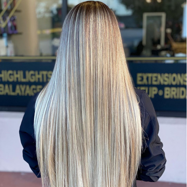 Back view of long straight blonde balayage in front of salon signage reading HIGHLIGHTS, BALAYAGE and EXTENSIONS