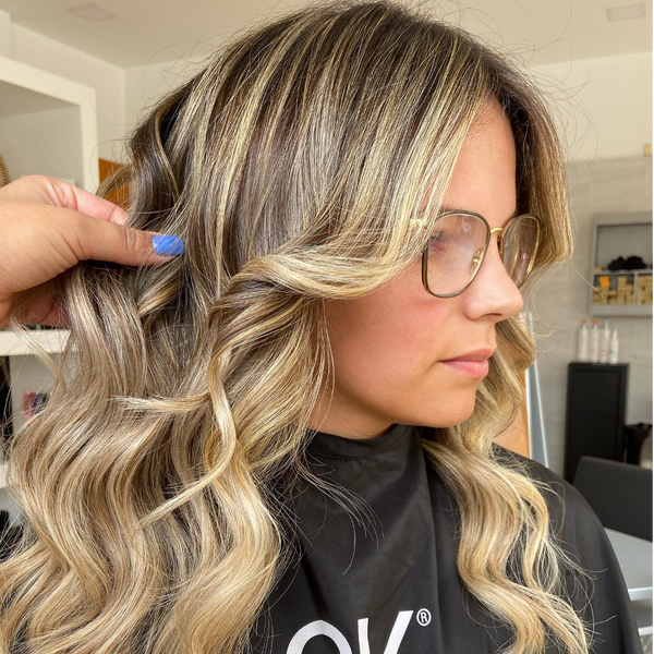 Side view of a woman in glasses in a black GK cape, a stylist lifting her waved blonde-highlighted hair