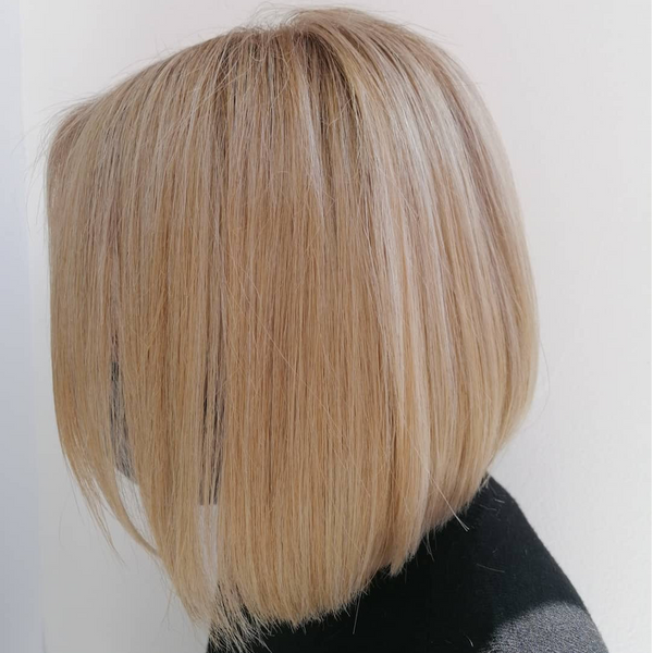 Blunt shoulder-length blonde bob seen from behind, sliced pale highlights and softly grown-out darker roots, on a white wall