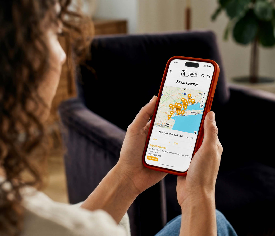 Woman with curly hair sitting in an armchair, holding a phone showing the GK Hair Salon Locator map