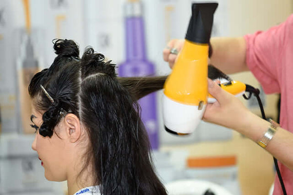 Salon stylist blow-drying a client's sectioned damp dark hair with a yellow dryer, blurred product shelves behind