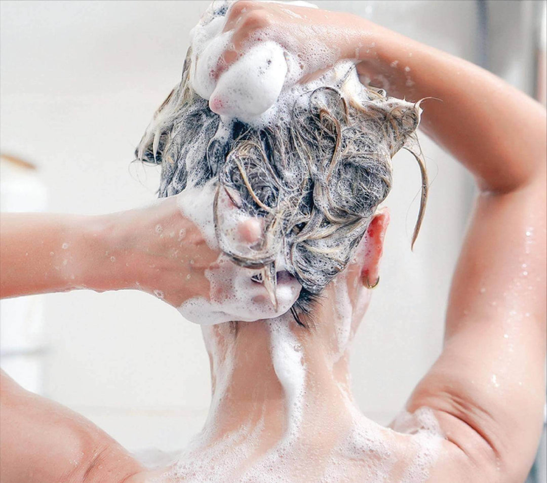 Seen from behind, a woman in the shower works thick white lather through her wet dark blonde hair with both hands